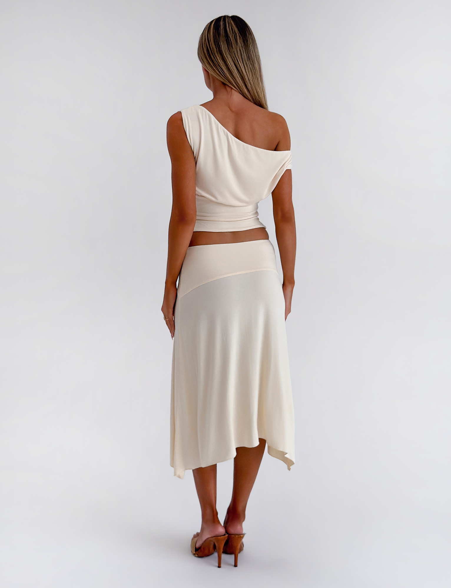 A woman stands facing away, wearing the TIGERMIST ALYSSA HI LO SKIRT in cream with an asymmetrical hem and front leg slit, paired with a matching cream one-shoulder top and heeled sandals against a plain white background.