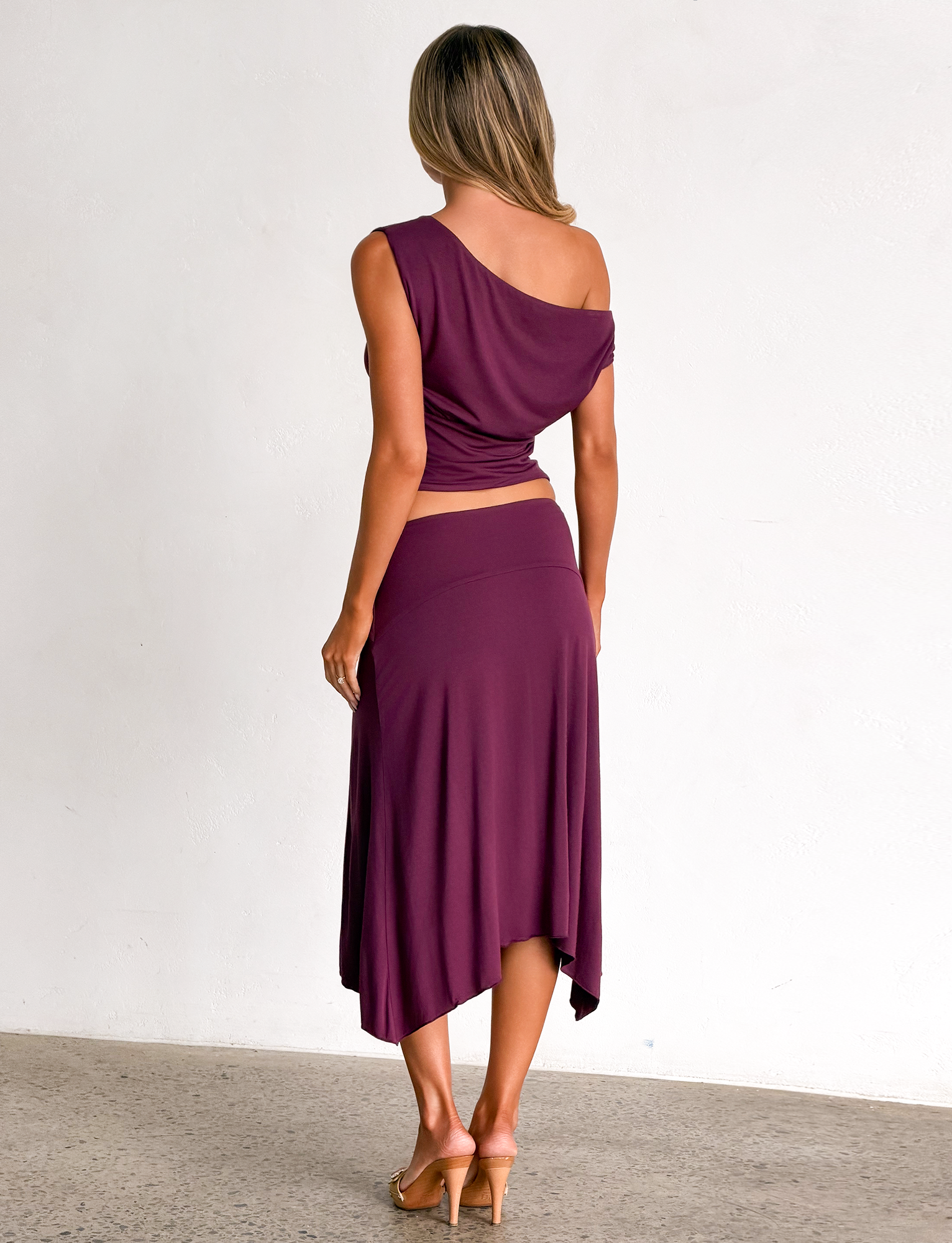 A woman stands indoors facing away, wearing the TIGERMIST ALYSSA OFF SHOULDER TOP in plum with a matching handkerchief-hem skirt and tan heeled sandals, against a plain white wall and concrete floor.