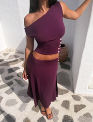 A woman wears the TIGERMIST ALYSSA OFF SHOULDER TOP in plum, a purple two-piece with white buttons, standing on a sunlit stone patio by a white wall, one arm raised and her head out of frame.