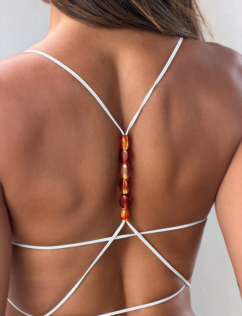 A close-up shows a woman wearing the TIGERMIST MINERVA 2.0 BODYSUIT in cream lace, featuring delicate vertical amber bead detailing along the back and a chic G-string bottom, set against a neutral background that highlights her tanned skin.