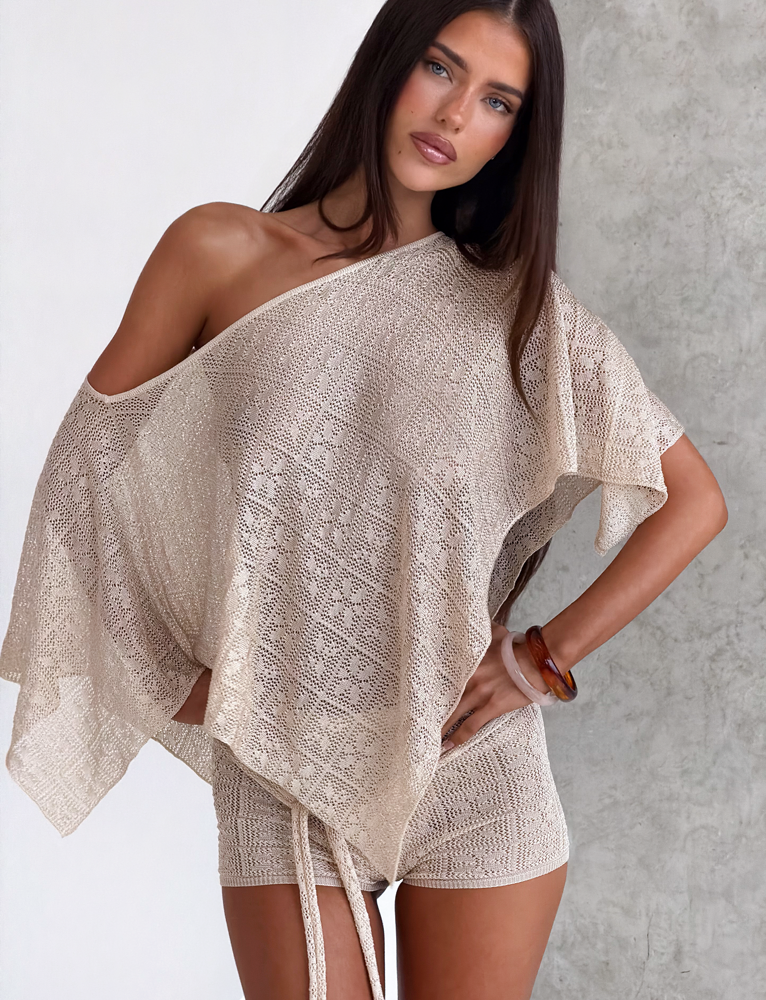 A woman with long dark hair wears the TIGERMIST FERN CROCHET PONCHO in Champagne, styled as an effortless beach cover-up, standing against a white and gray textured wall and gazing neutrally at the camera.