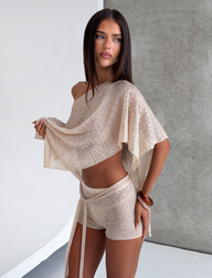 A woman with long brown hair poses by a textured gray wall, wearing the CL - FERN CROCHET SHORT from Tiger Mist USA with a matching sheer, off-shoulder top, holding up her top and gazing to the side neutrally.