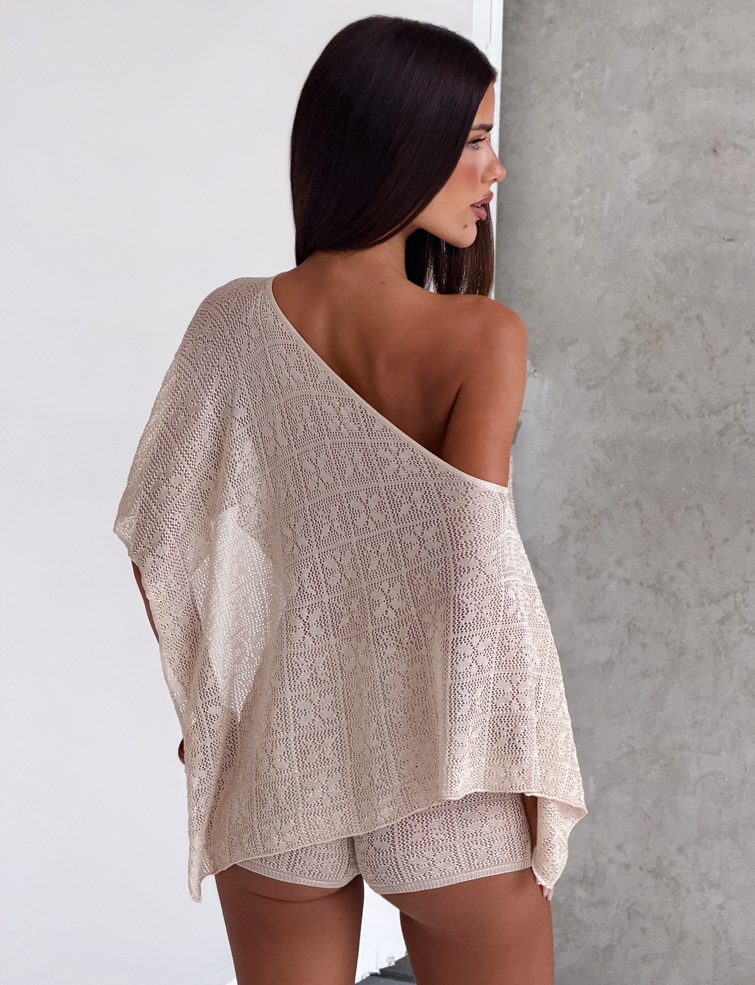 A woman with long dark hair stands sideways, wearing TIGERMIST’s FERN CROCHET SHORT in Champagne with a matching semi-sheer one-shoulder knitted top. She faces a textured gray wall, her shoulder and back exposed.