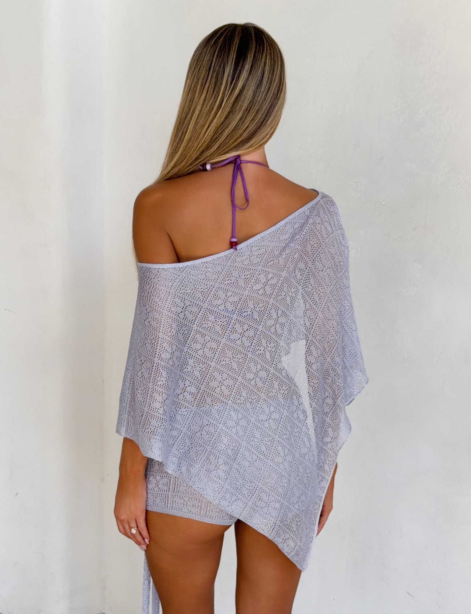 A woman with long, straight hair stands facing away in the TIGERMIST FERN CROCHET PONCHO in silver over a purple halter bikini, set against a plain white wall.