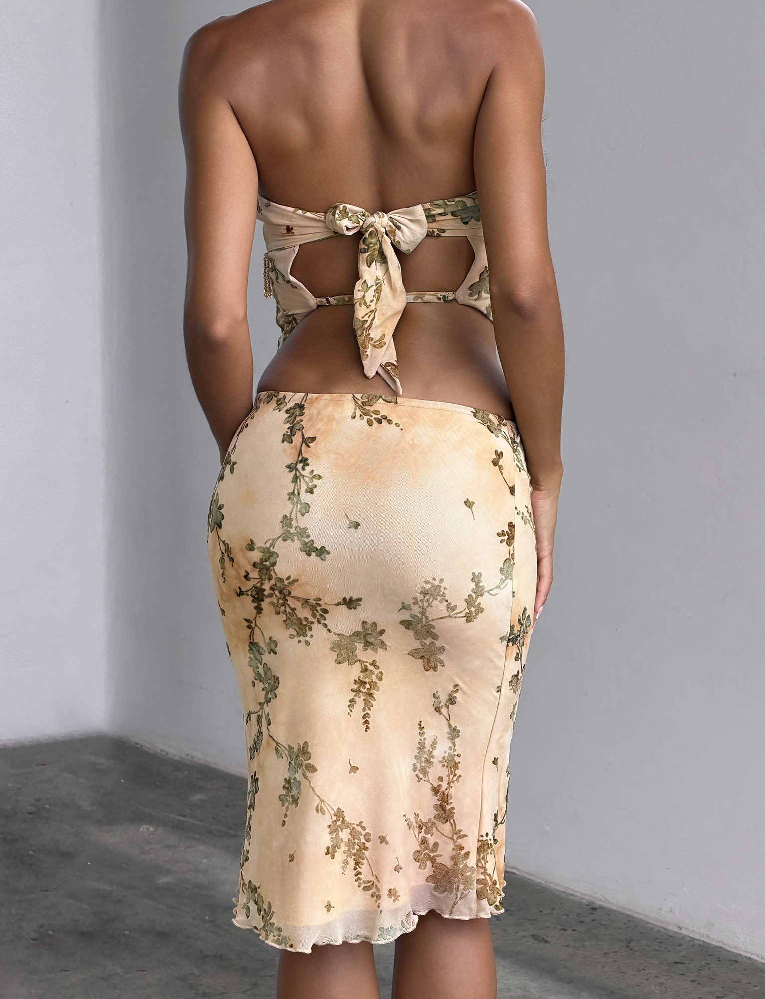 A woman stands with her back to the camera, wearing the OONA BURN OUT FRINGE SKIRT - TAN by TIGERMIST, a fitted silhouette featuring tan fringe detailing and a removable waistband for versatile styling.