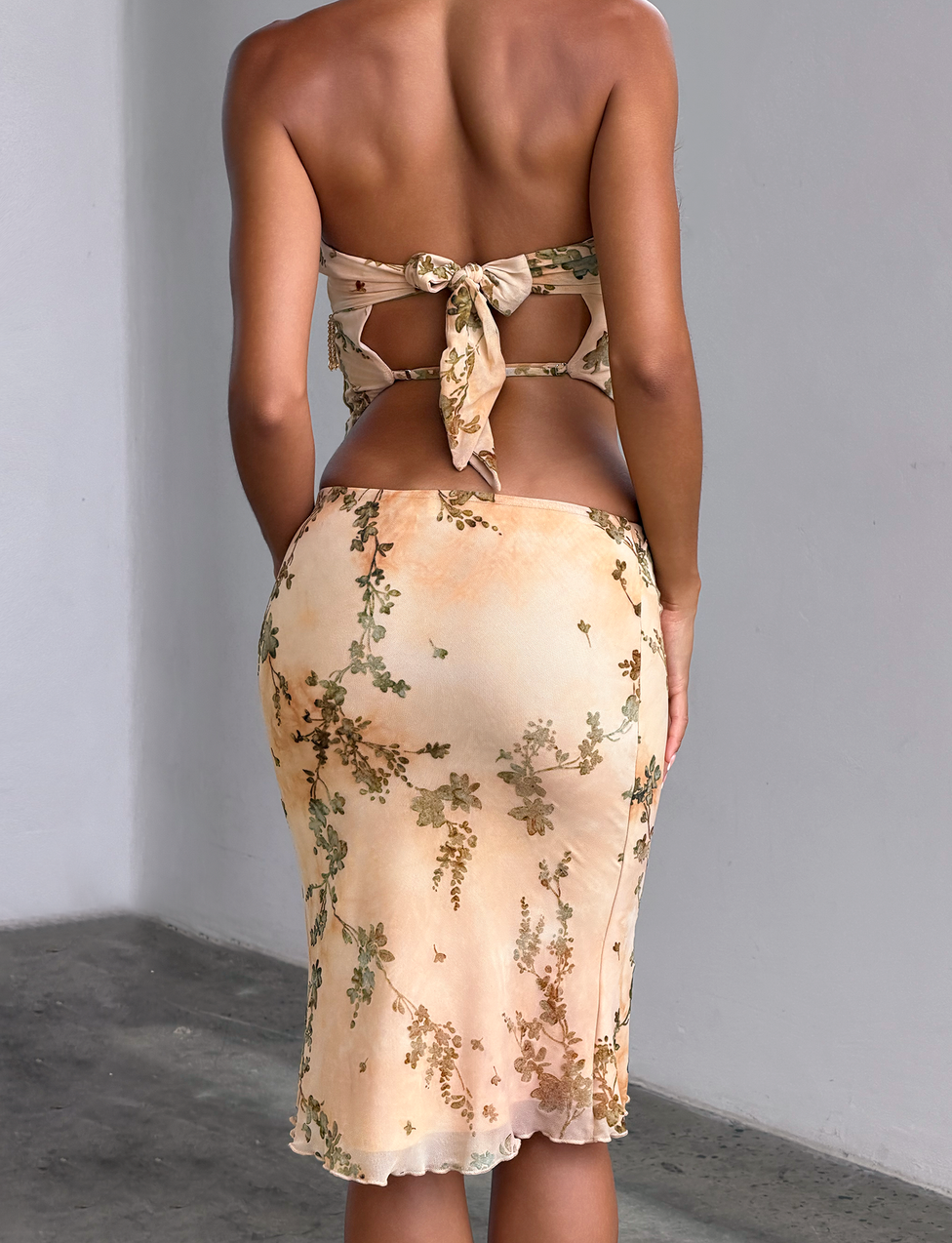 A woman stands with her back to the camera, wearing the OONA BURN OUT FRINGE SKIRT - TAN by TIGERMIST, a fitted silhouette featuring tan fringe detailing and a removable waistband for versatile styling.