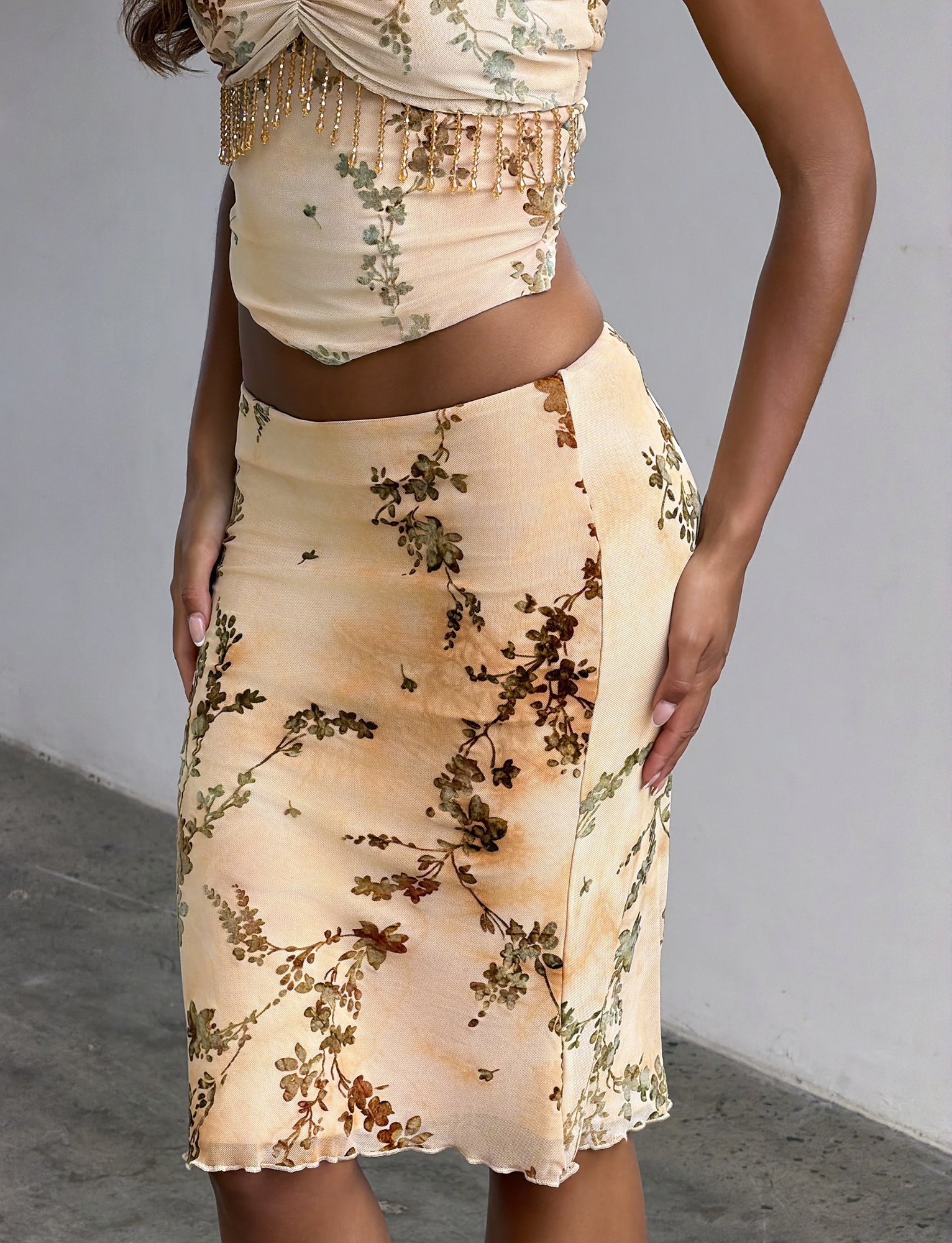 A woman stands by a light-colored wall wearing the TIGERMIST OONA BURN OUT FRINGE SKIRT in tan, paired with a matching sleeveless crop top featuring bead details.