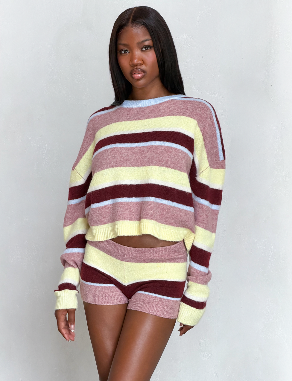 A woman wearing the TIGERMIST KOA 2.0 STRIPED JUMPER in tan stripe stands against a light gray background, gazing at the camera with a neutral expression.