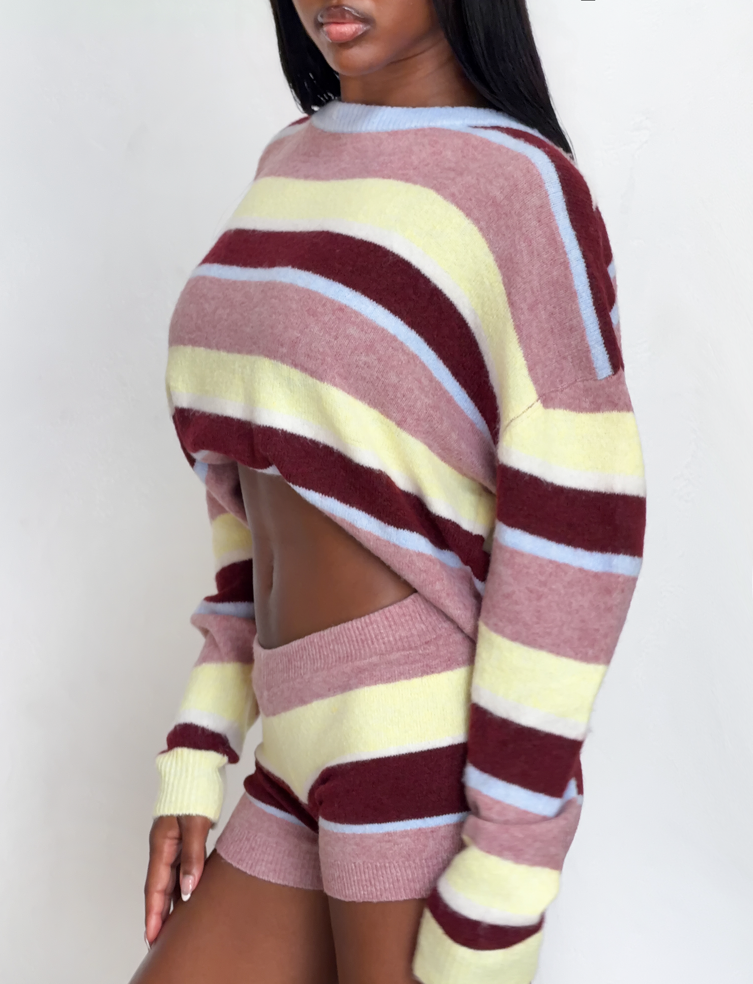 A woman wears TIGERMIST's KOA 2.0 STRIPED SHORT in tan stripe and a coordinating long-sleeve sweater with maroon, yellow, and light blue stripes, standing against a plain white background.