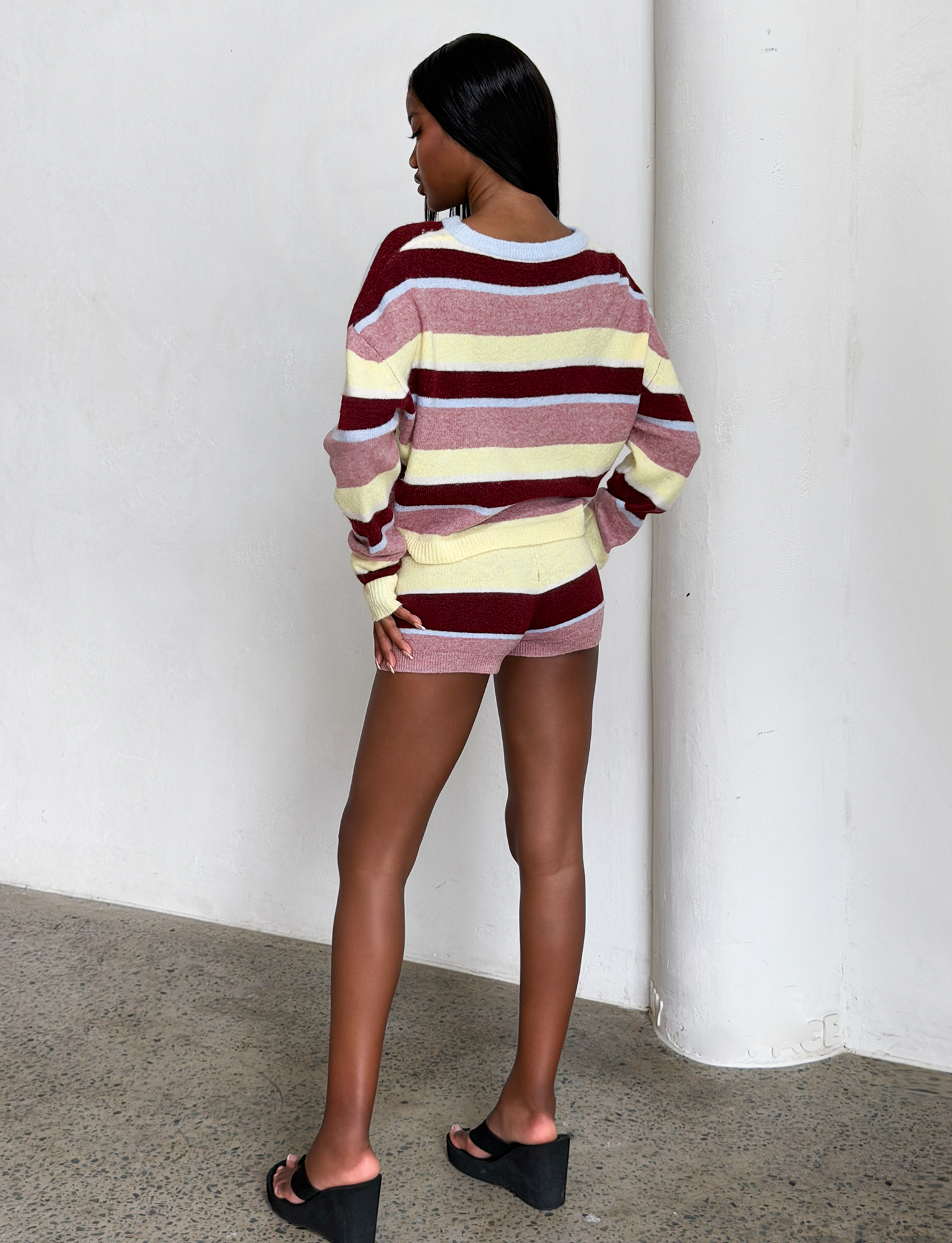 A woman stands indoors facing away, wearing the TIGERMIST KOA 2.0 STRIPED JUMPER in tan stripe, paired with matching shorts, and black platform sandals on a concrete floor.
