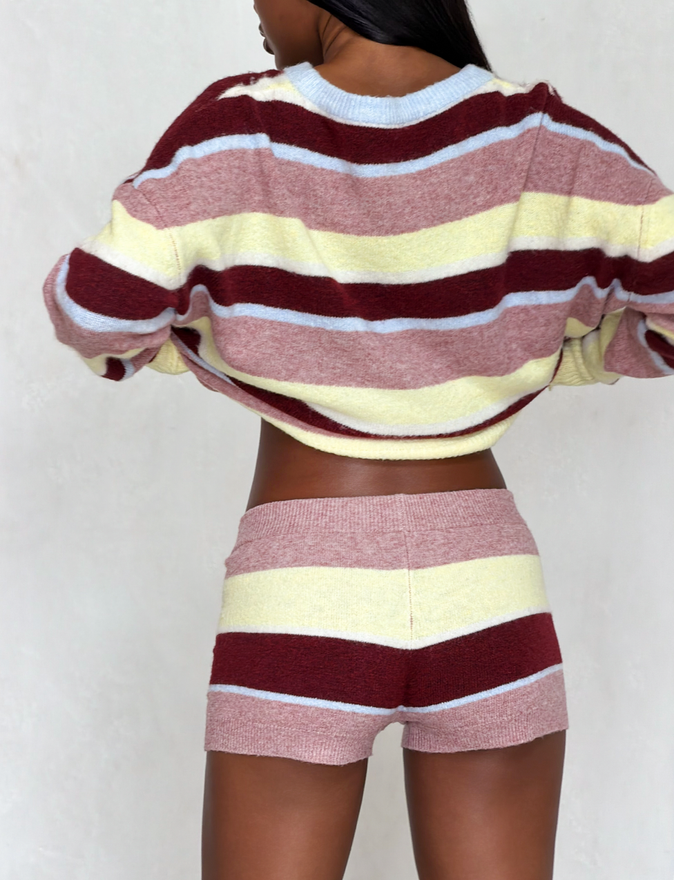 A woman faces away from the camera, wearing a cozy knit sweater and TIGERMIST KOA 2.0 STRIPED SHORT - TAN STRIPE with multicolour maroon, yellow, and light blue stripes for a relaxed, comfortable look.