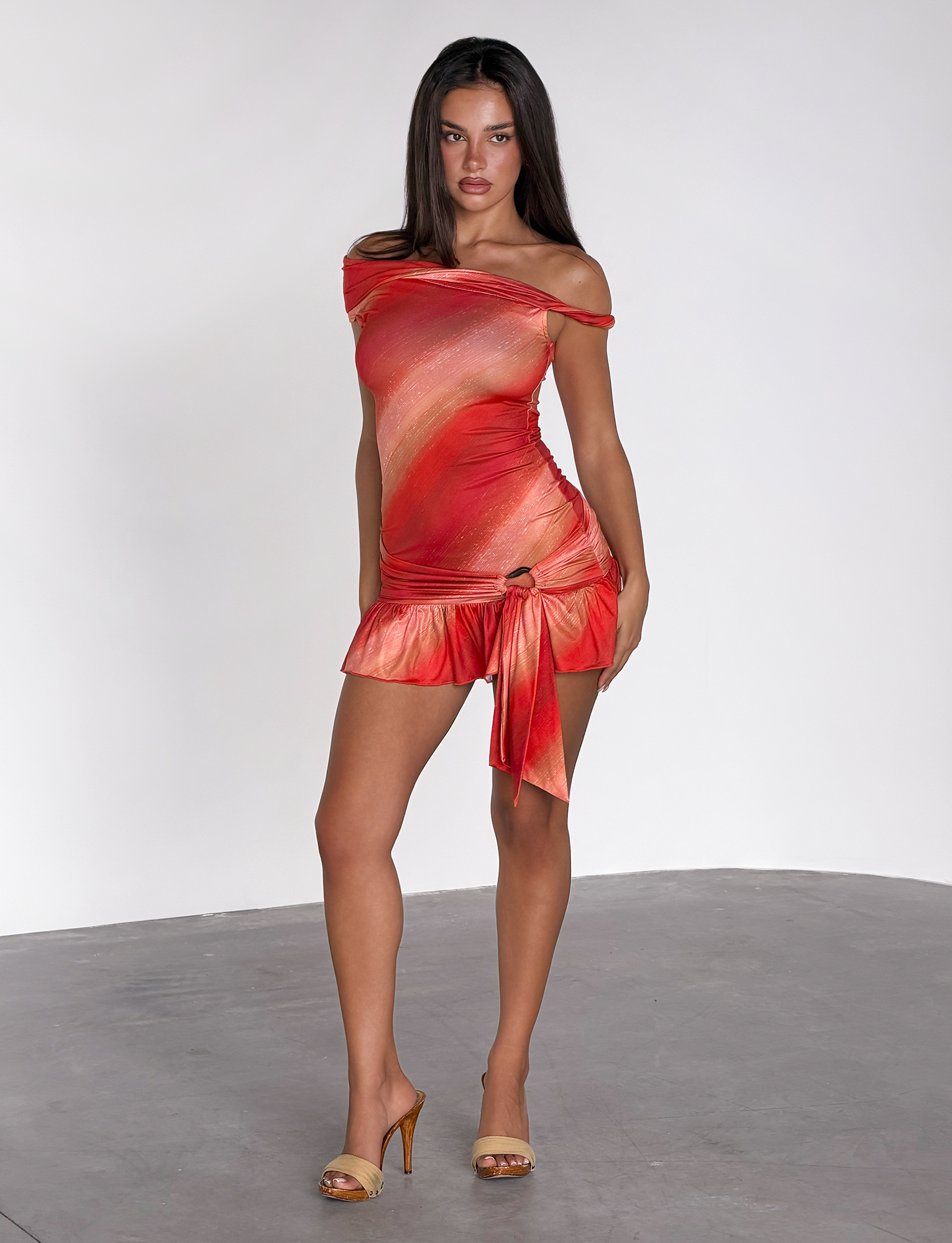 A woman stands confidently in the TIGERMIST HAELO MINI DRESS – SUNRISE GLITTER STRIPE, a sleeveless orange and red ombre dress with a tie detail and asymmetrical neckline, paired with tan heels against a plain white background.