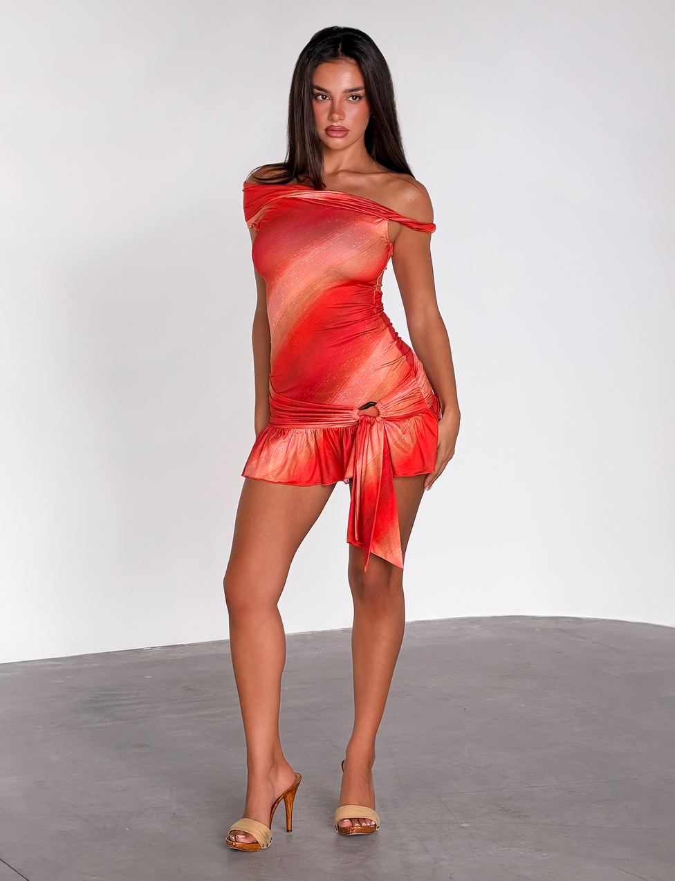 A woman stands confidently in the TIGERMIST HAELO MINI DRESS – SUNRISE GLITTER STRIPE, a sleeveless orange and red ombre dress with a tie detail and asymmetrical neckline, paired with tan heels against a plain white background.