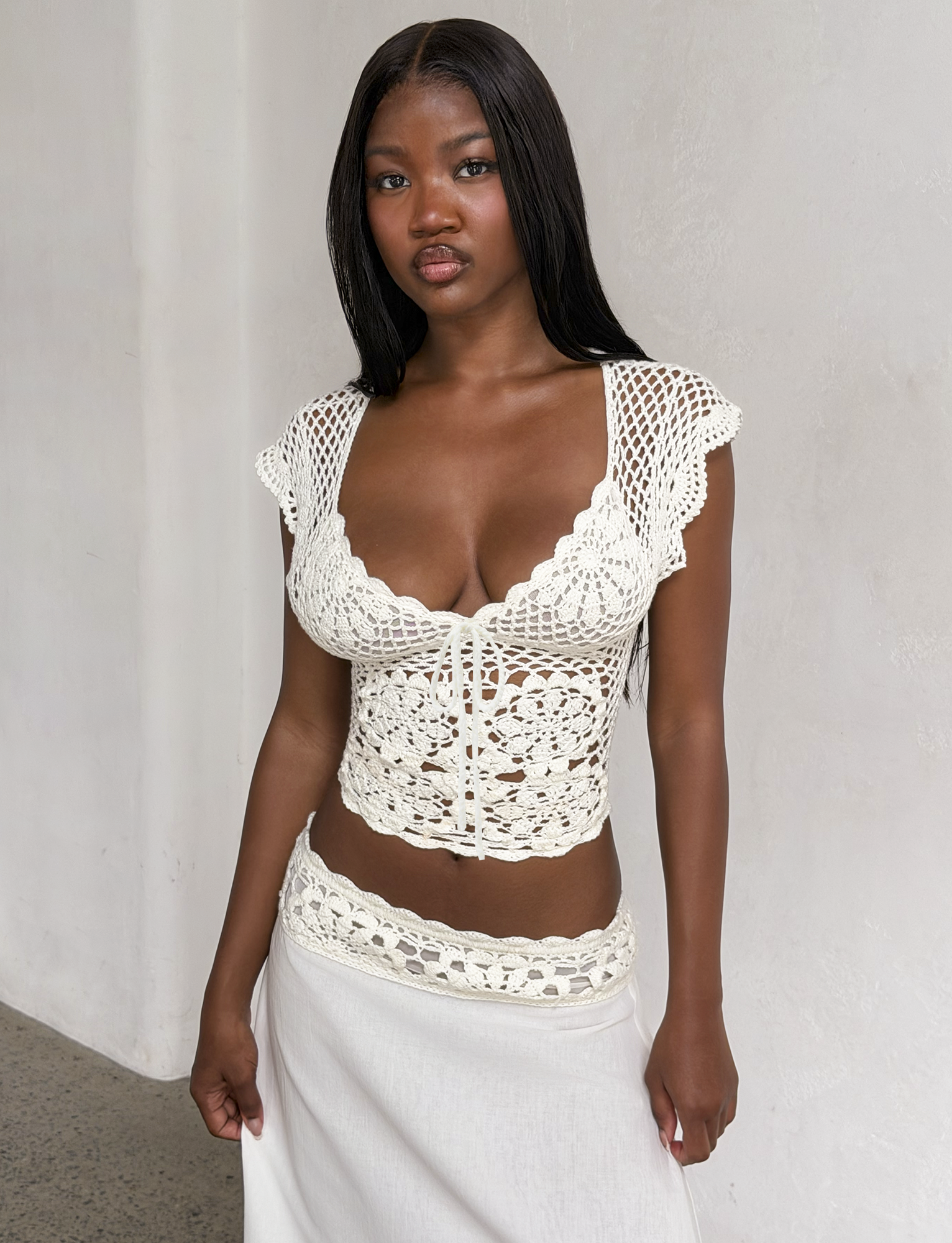 A woman with long black hair wears the TIGERMIST SASHA CROCHET TOP in ivory, featuring scalloped cap sleeves, as she stands against a plain white wall and looks at the camera.