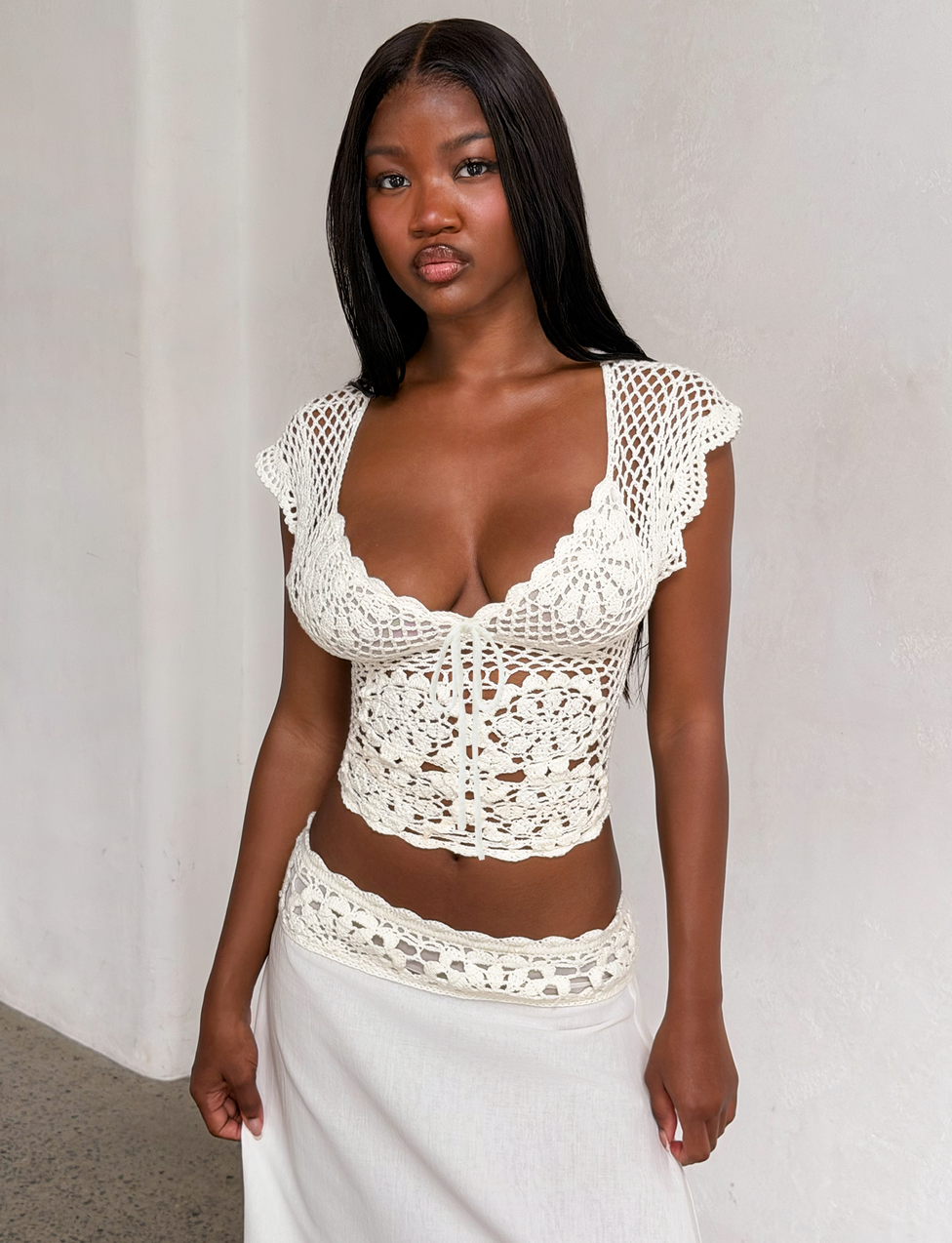 A woman with long black hair wears the TIGERMIST SASHA CROCHET TOP in ivory, featuring scalloped cap sleeves, as she stands against a plain white wall and looks at the camera.