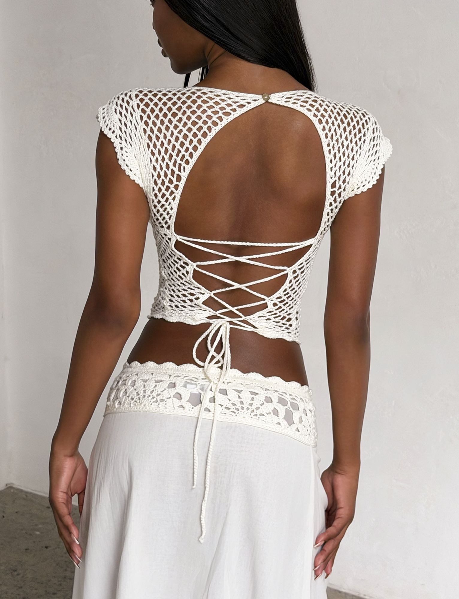 A woman wears the SASHA CROCHET TOP in ivory by TIGERMIST, featuring scalloped cap sleeves and crisscross ties, paired with a flowing white skirt. She stands indoors, facing away from the camera.