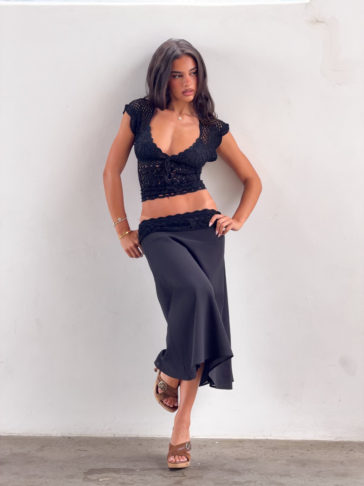 A woman with long dark hair wears the TIGERMIST SASHA CROCHET TOP in black, featuring a lace-up back, paired with a long black skirt. She leans against a white wall, finishing her chic look with wedge sandals and minimal jewelry.