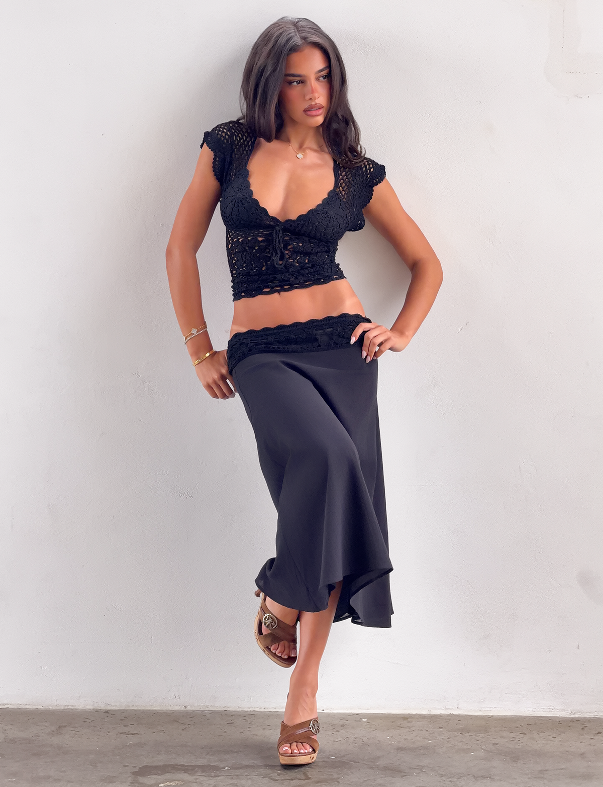 A woman with long dark hair stands against a light wall, wearing the TIGERMIST SASHA CROCHET MIDI SKIRT in black, a matching crochet top, brown heels, and gold jewelry. She looks to the side with one hand on her hip.