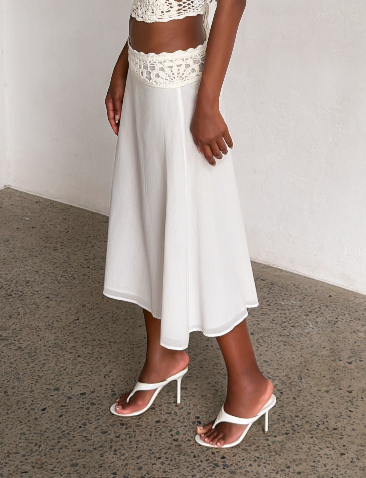 Wearing a white crochet crop top, TIGERMIST SASHA CROCHET MIDI SKIRT - IVORY, and white high-heeled sandals, a person stands on a speckled concrete floor against a white wall.