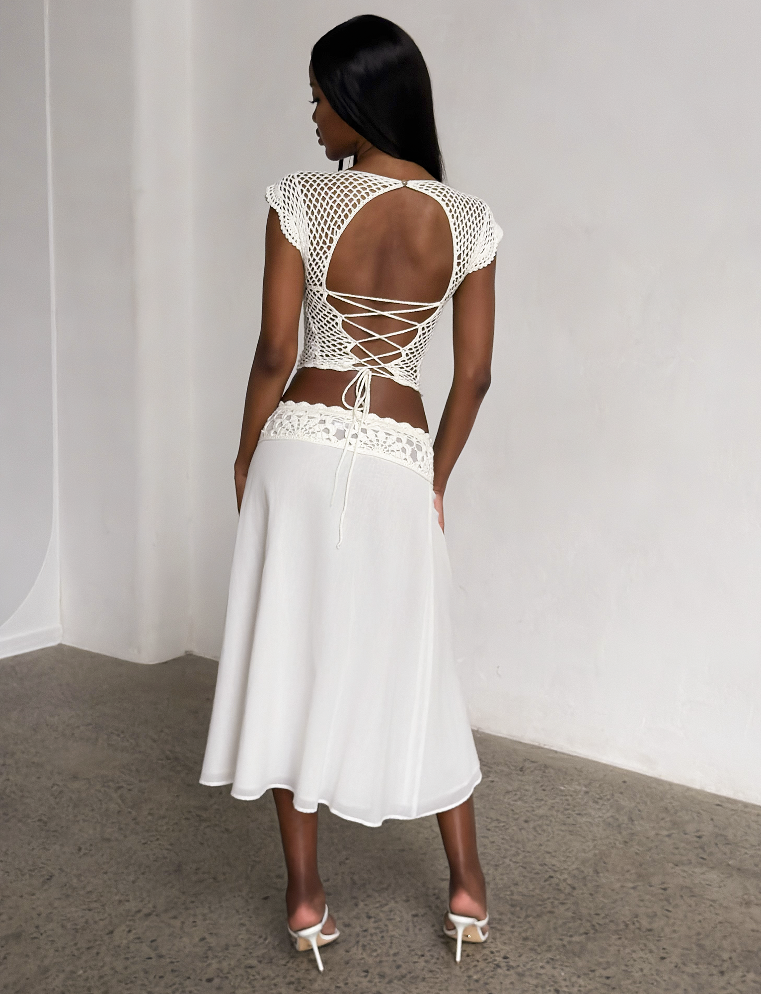 A woman stands indoors facing away, wearing a white crocheted lace top with an open back and crisscross ties, paired with the TIGERMIST SASHA MIDI SKIRT in ivory and heeled sandals. The background is minimal and neutral.