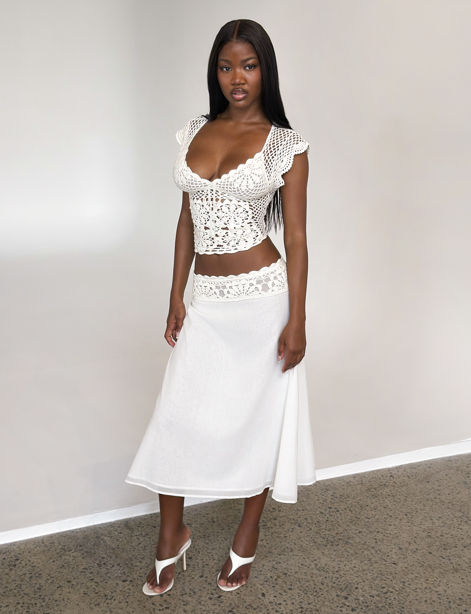 A woman stands indoors against a plain background, wearing the TIGERMIST SASHA MIDI SKIRT in ivory, paired with a white crochet cap-sleeve top and white heeled sandals.