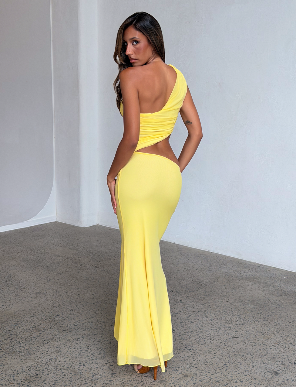 A woman models the TIGERMIST COBI 2.0 MAXI DRESS in butter yellow, featuring a one-shoulder neckline and asymmetrical design, posing with her back turned on a polished concrete floor in a minimalist, light-filled room.