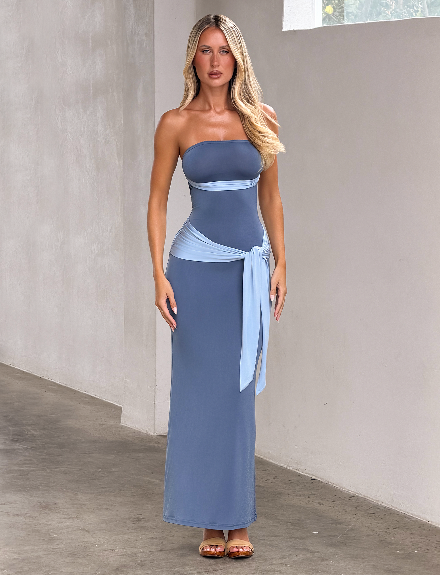 A woman poses indoors against a light wall, wearing the TIGERMIST TOVE MAXI DRESS in blue—a strapless, fitted silhouette with a light blue waist sash. She has long blonde hair.