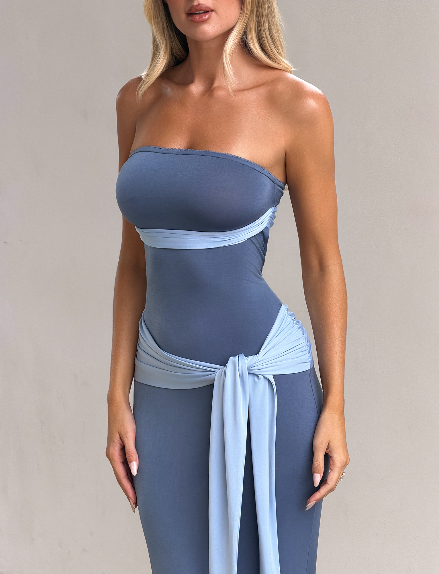 A woman models the TIGERMIST TOVE MAXI DRESS - BLUE, featuring a strapless, fitted silhouette with a light blue sash at the waist, standing against a plain light background. Her face is not visible.
