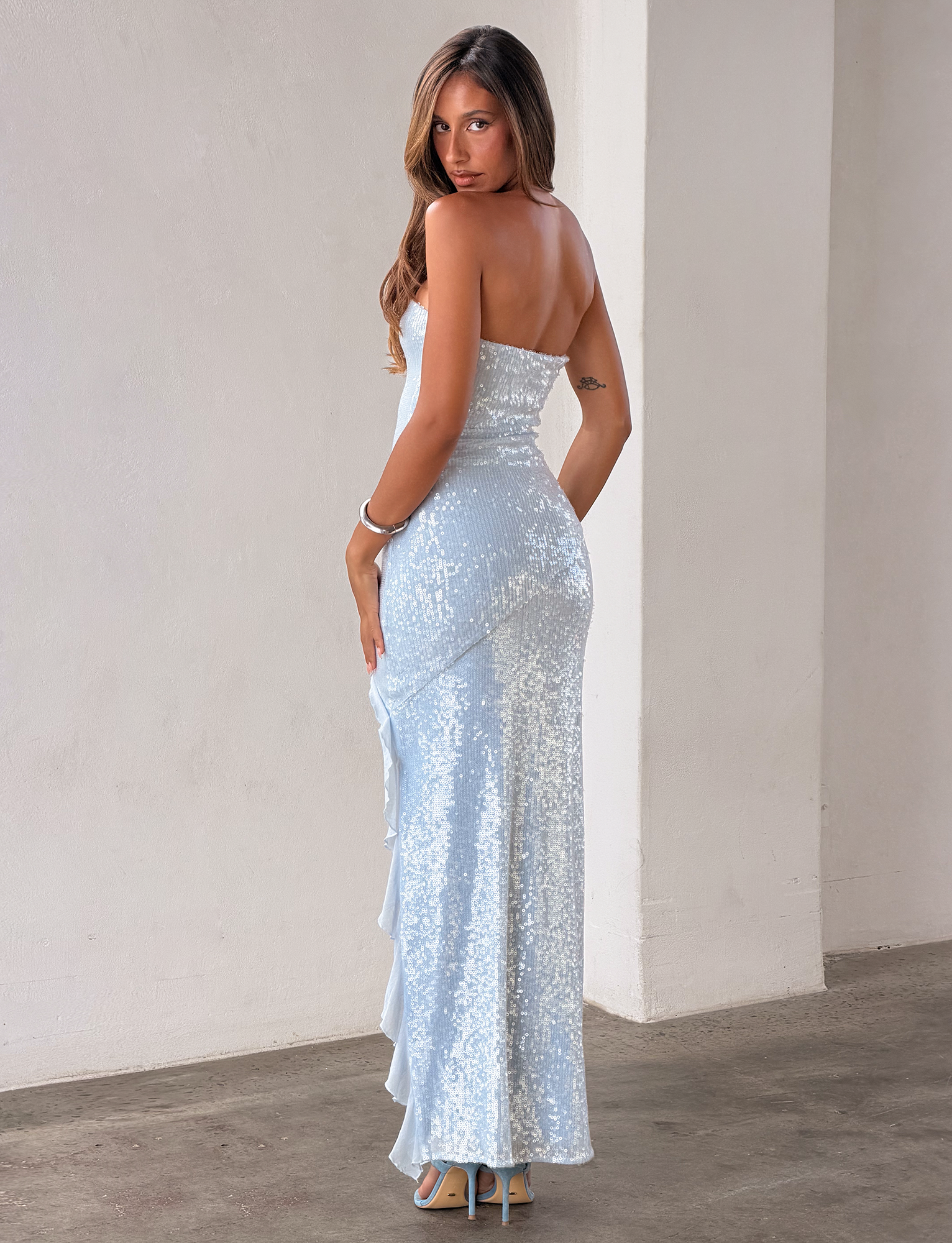 A woman in the TIGERMIST THEO SEQUIN MAXI DRESS - SOFT BLUE, featuring a mermaid silhouette, poses on a concrete floor with long hair and high heels in a minimalist, light-toned setting, turning her head toward the camera.