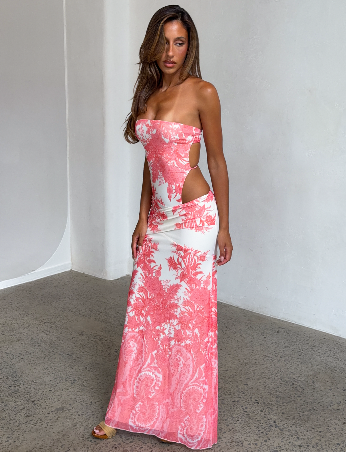 A woman stands indoors near a white wall, wearing the TIGERMIST POPPI MAXI DRESS - CORAL TAPESTRY featuring a strapless neckline and cutout mesh design with pink floral patterns.