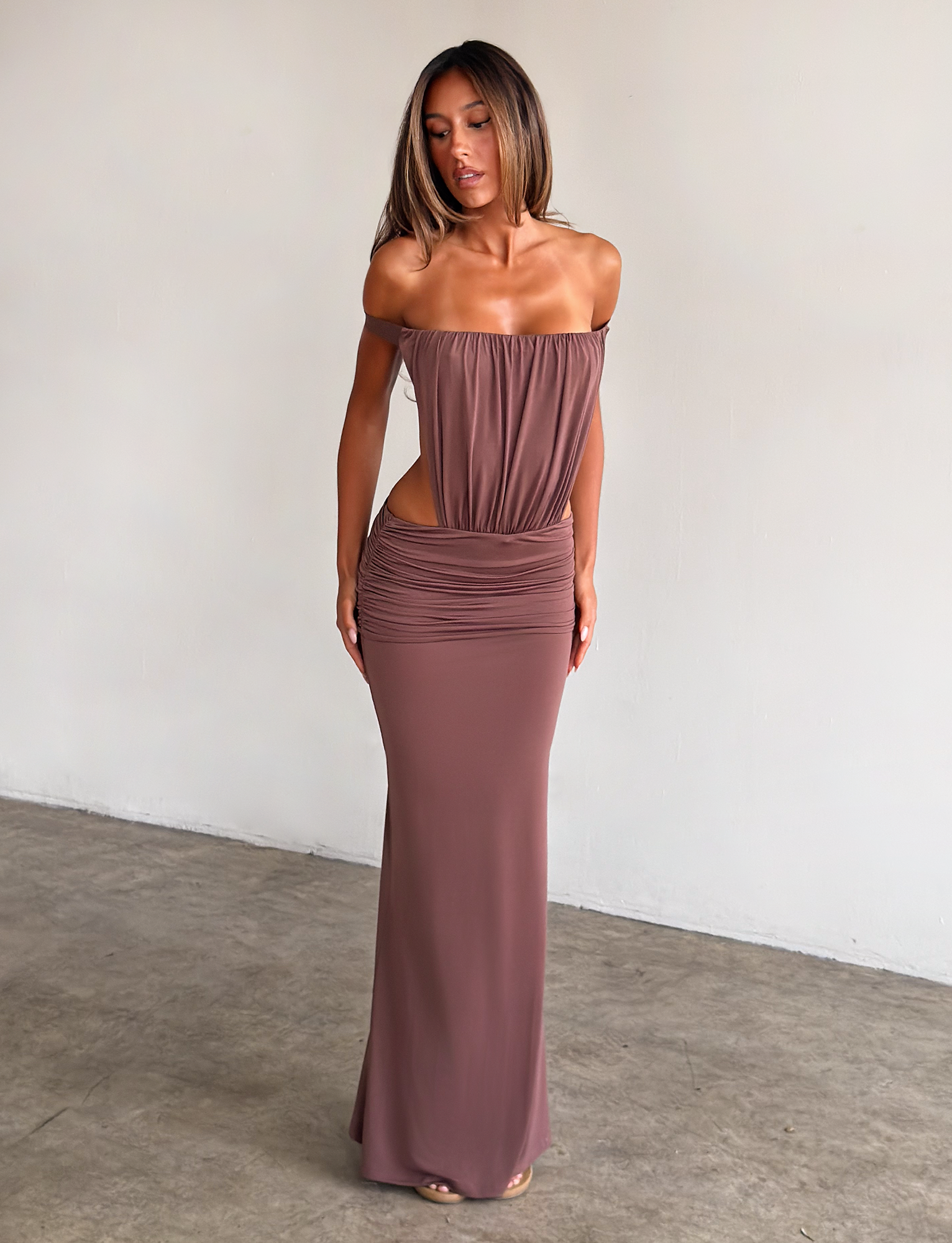 A woman stands indoors wearing the TIGERMIST IOLLA STRAPLESS MAXI DRESS in mocha, featuring a ruched bodice, cut-out sides, and a long fitted skirt. She gazes down with straight hair and neutral makeup against a plain light background.