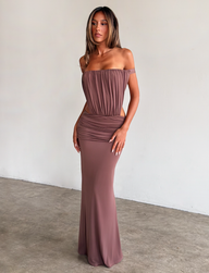 A woman stands against a plain light wall, elegantly posing on concrete in the TIGERMIST IOLLA STRAPLESS MAXI DRESS in mocha, featuring an off-the-shoulder ruched bodice, side cutouts, and waist detailing.