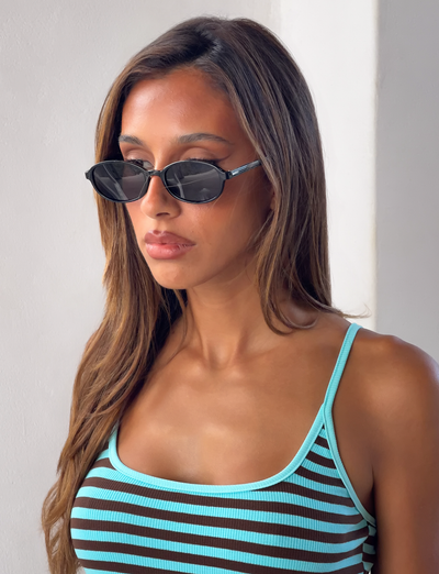 A woman with long brown hair wears Tiger Mist USA CL - ESTA SUNGLASSES and a turquoise-striped tank top, standing against a white wall and looking to the side with a neutral expression.