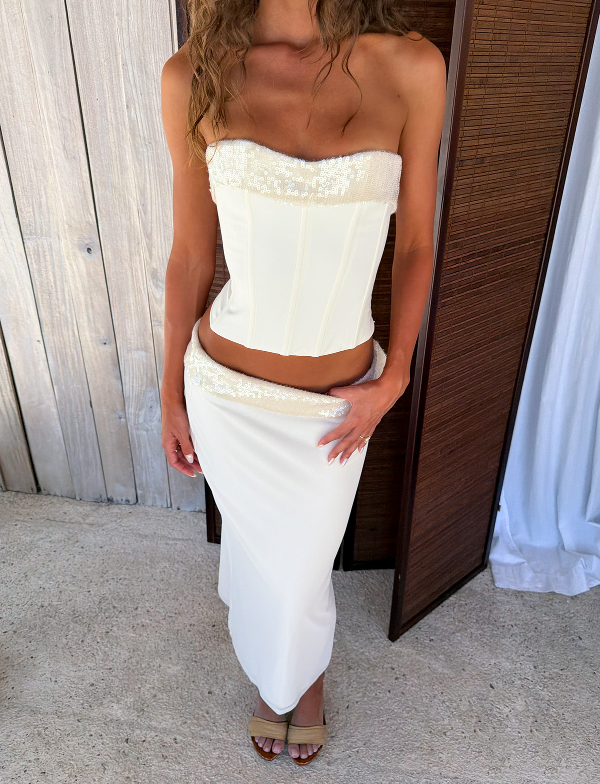A woman wears the TIGERMIST CHARLYZE BUSTIER in cream—a strapless corset top with sequin trim and lace-up back—paired with a matching low-rise maxi skirt featuring a sequin waistband and nude heeled sandals.