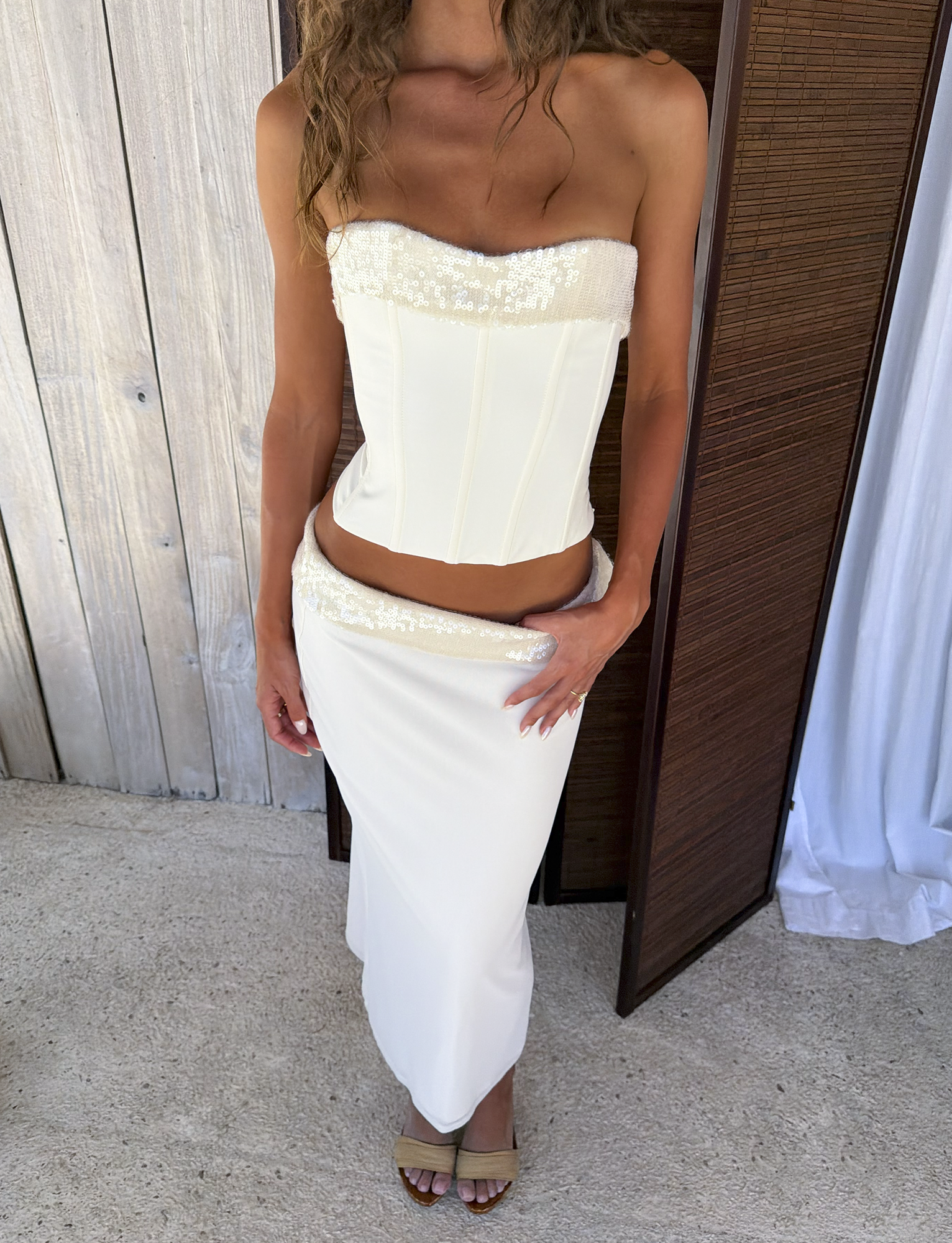 A woman wears the TIGERMIST CHARLYZE BUSTIER in cream—a strapless corset top with sequin trim and lace-up back—paired with a matching low-rise maxi skirt featuring a sequin waistband and nude heeled sandals.