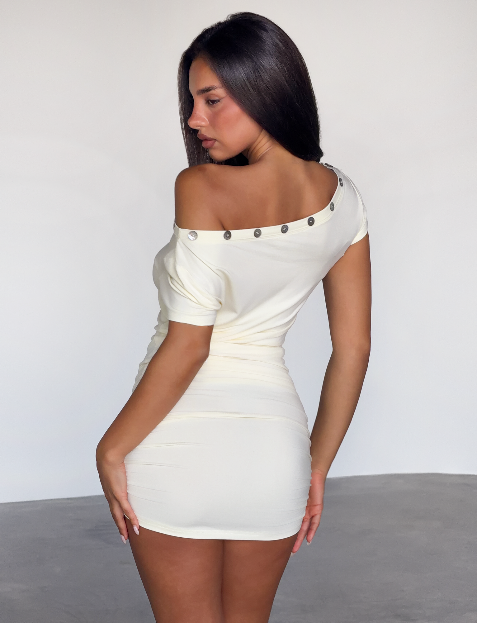 A woman with long dark hair poses in a fitted, off-the-shoulder TIGERMIST MARITA MINI DRESS - CREAM, featuring metallic button details along the asymmetrical neckline, standing on a smooth gray floor against a plain light background.