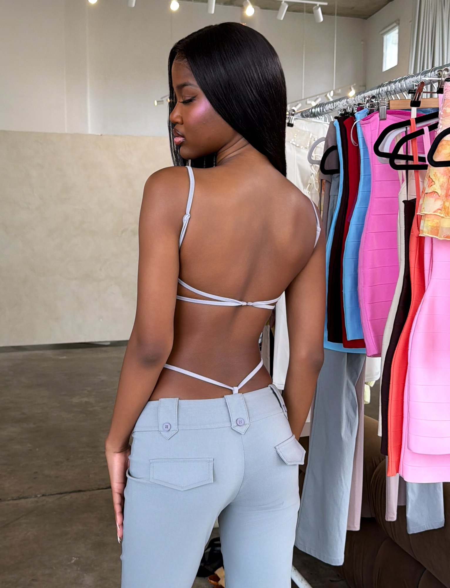 A woman faces away from the camera in a modern room with clothing racks, wearing TIGERMIST's MINERVA BODYSUIT in Frost Grey and light pants.