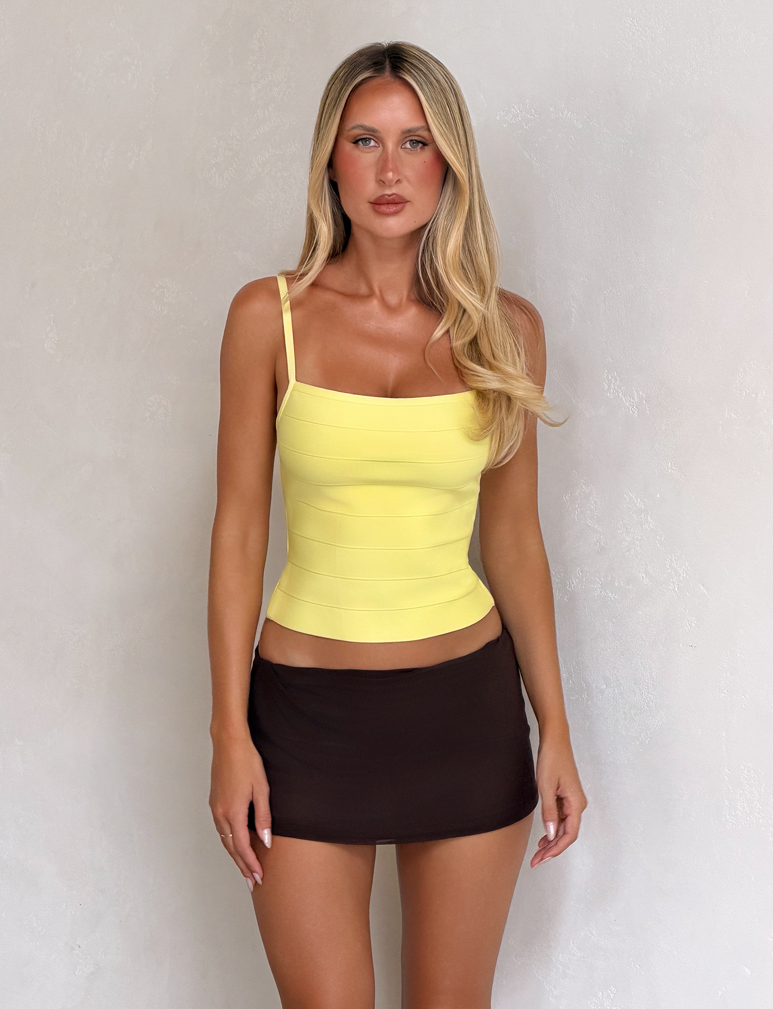 A woman with long blonde hair stands against a light wall, wearing the TIGERMIST MIDORI BANDAGE TOP in yellow and a short brown skirt, looking directly at the camera with a neutral expression.