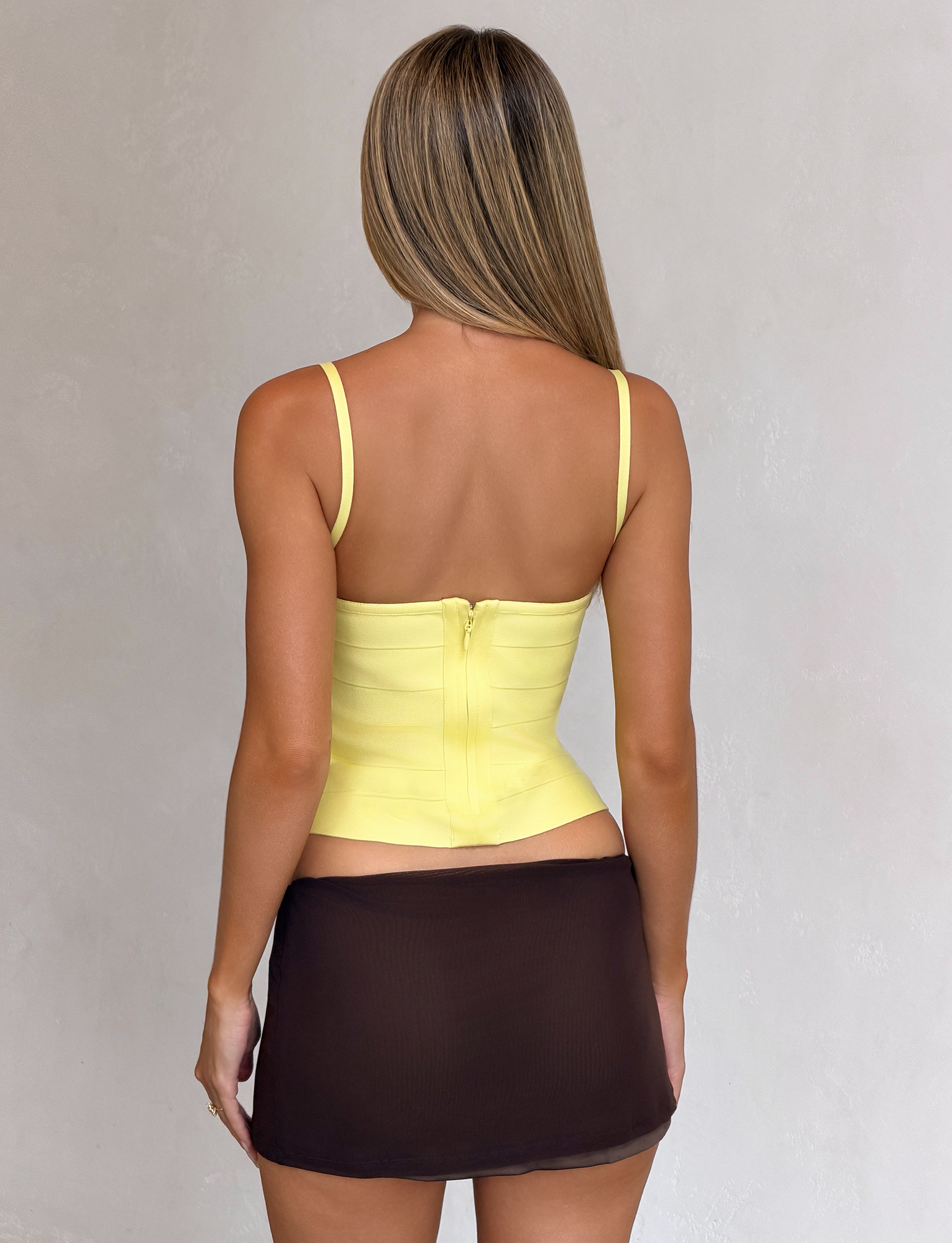 A woman with straight, light brown hair faces away, wearing the TIGERMIST MIDORI BANDAGE TOP in yellow with a back zipper and square neckline, paired with a short sheer black skirt against a plain light background.