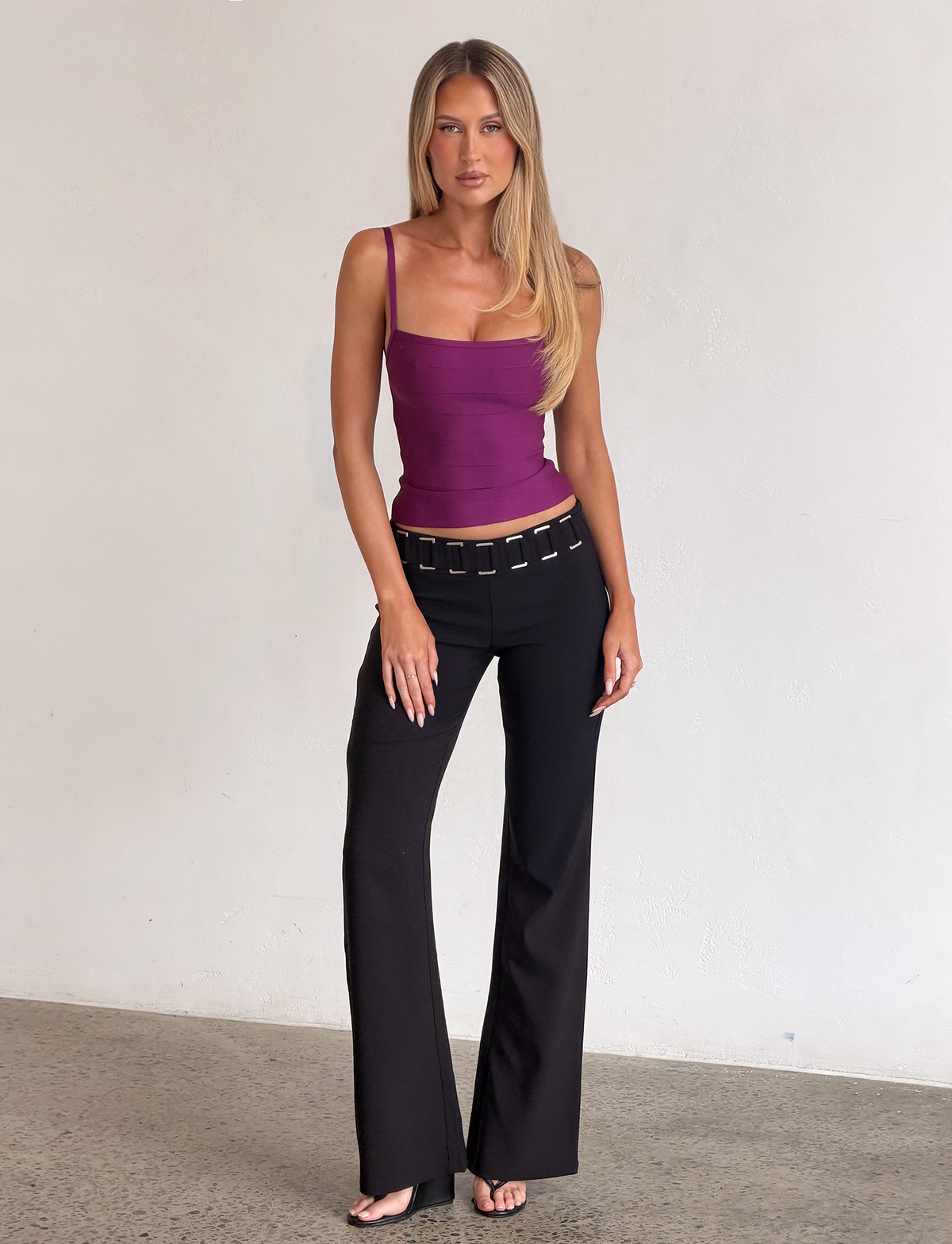 A woman with long blonde hair and a neutral expression stands against a plain white wall, wearing the TIGERMIST MIDORI BANDAGE TOP in plum—a square-neck tank that subtly cinches at the waist—paired with black flared pants featuring a chain belt and black heels.