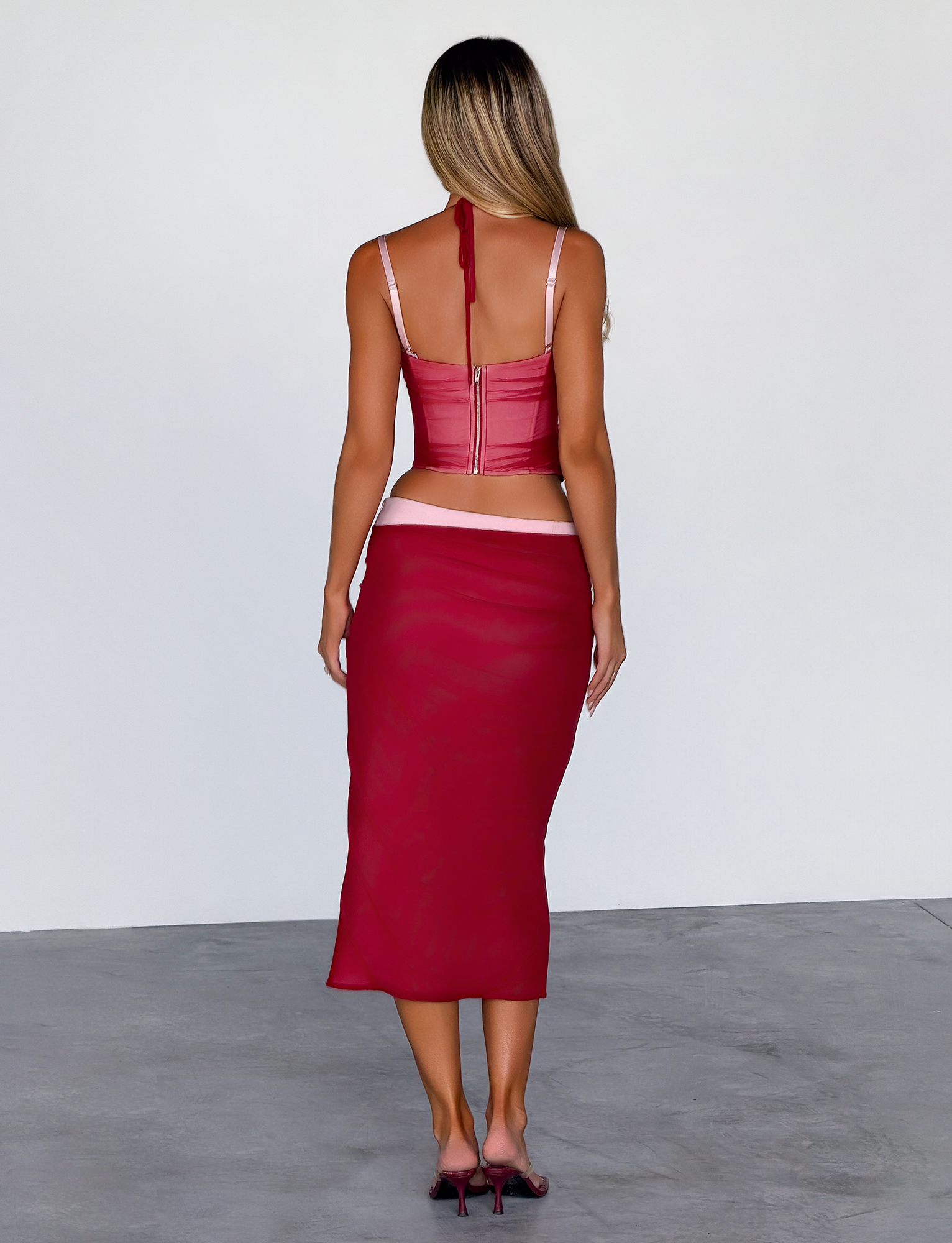 A woman with long, wavy blonde hair faces away, wearing the TIGERMIST CALINE MIDI SKIRT in red with a fitted crop top and matching heels, standing on gray concrete against a plain white wall.