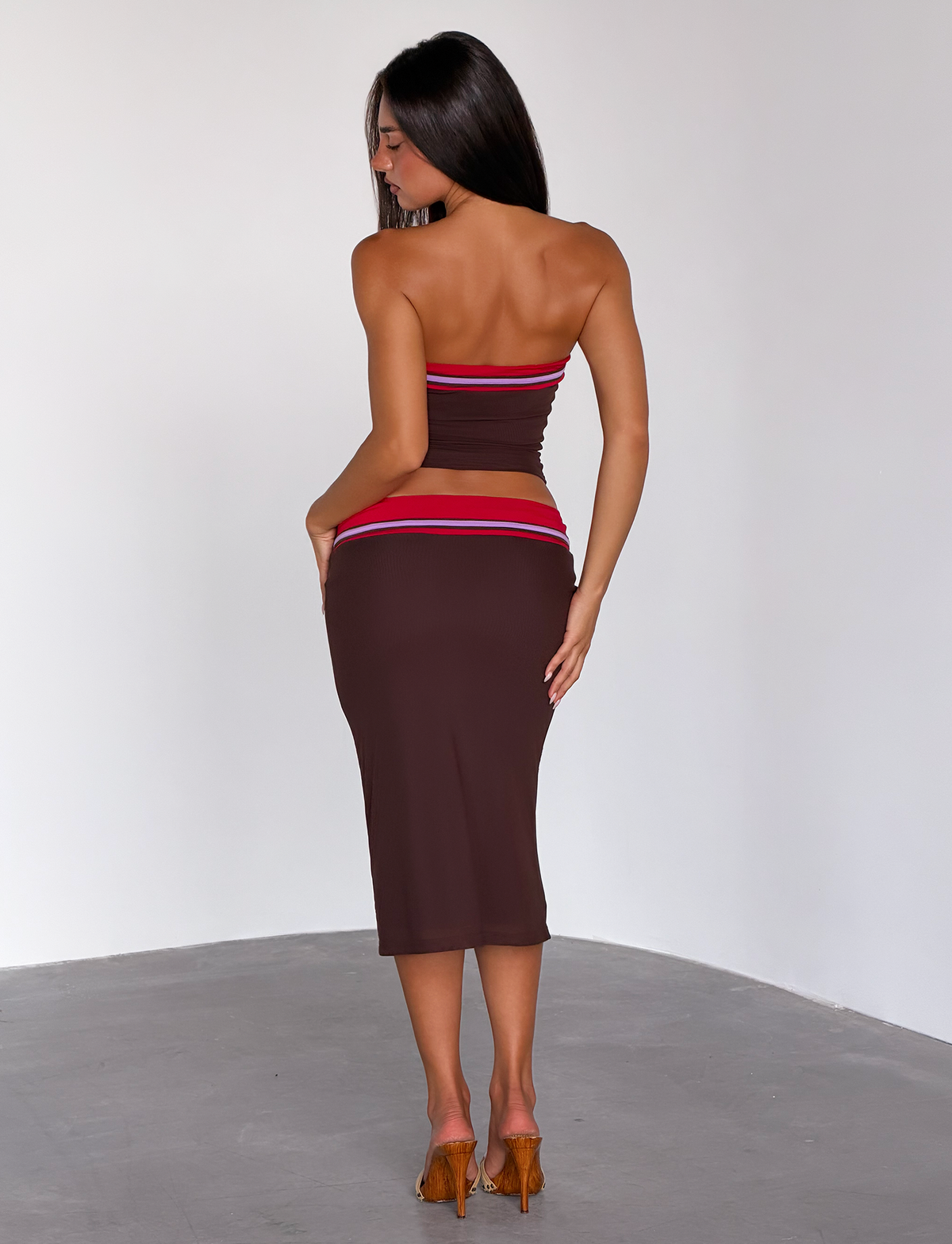 A woman with long dark hair stands indoors facing away, wearing TIGERMIST's SERAH DIAMANTE MIDI SKIRT in chocolate, paired with a 90's strapless brown top and brown heels. The room has white walls and a gray floor.