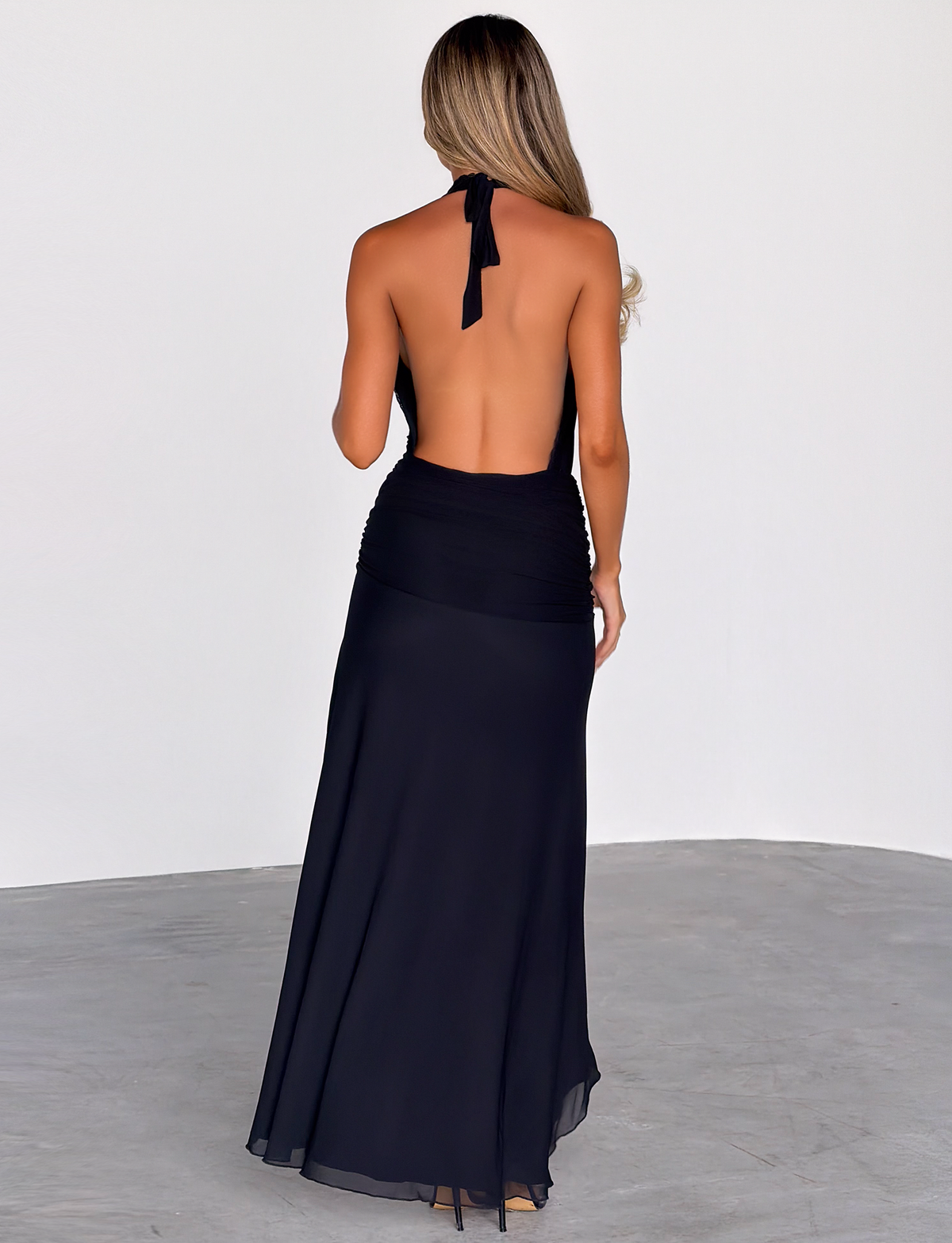 A woman with long, wavy hair stands with her back to the camera, wearing the TIGERMIST CORELLE DRESS in black—a floor-length plunge maxi—on a plain gray floor against a white wall background.
