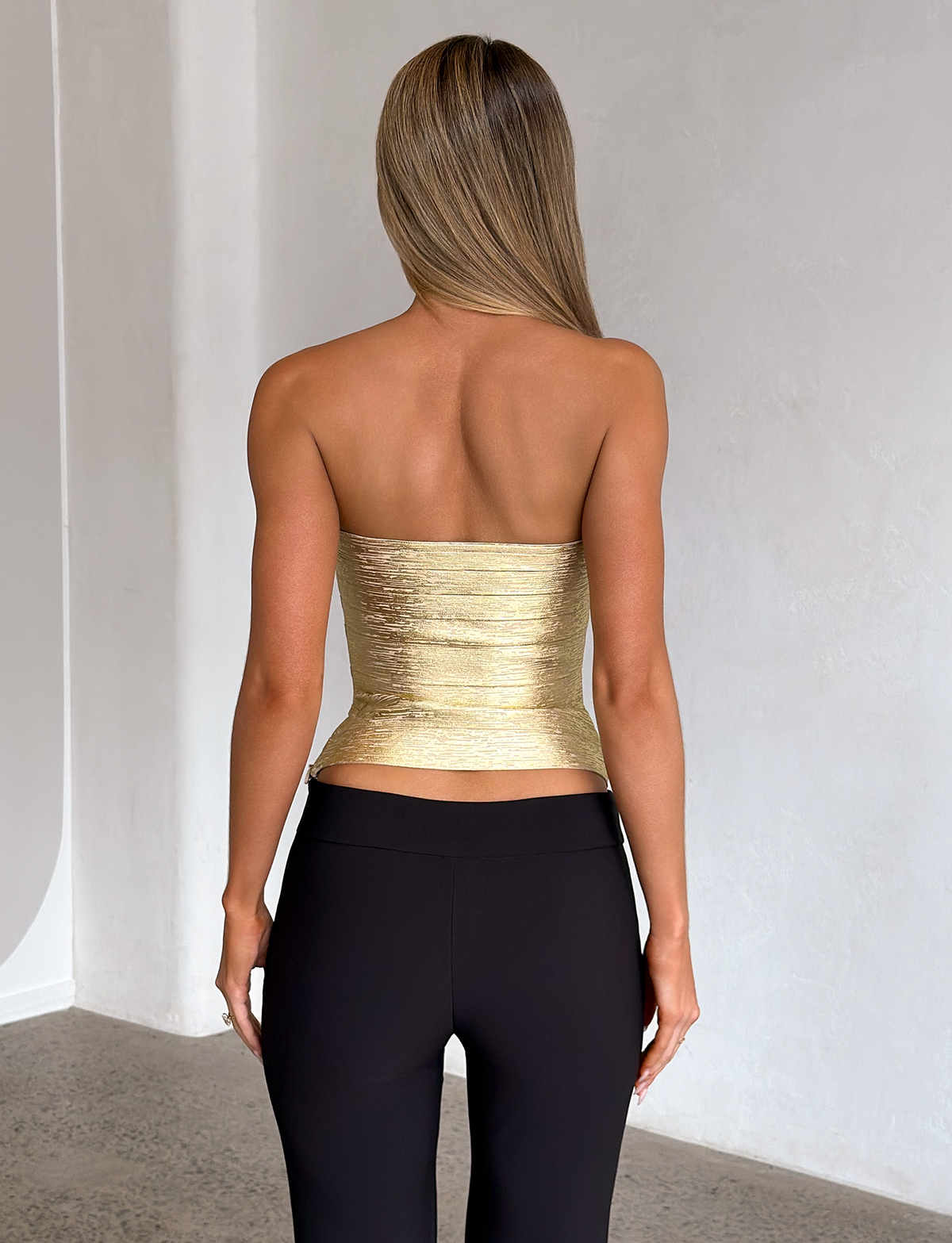 A woman with straight, blonde hair stands indoors facing away, wearing the TIGERMIST NIYA METALLIC BANDAGE TOP in gold paired with black pants, set against a light-colored wall and textured floor.