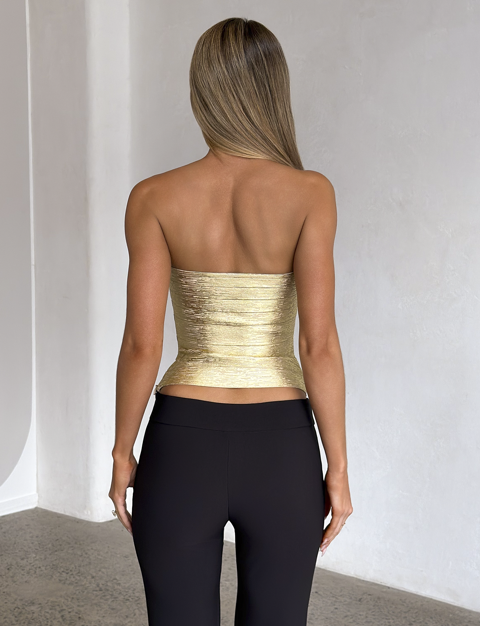 A woman with straight, blonde hair stands indoors facing away, wearing the TIGERMIST NIYA METALLIC BANDAGE TOP in gold paired with black pants, set against a light-colored wall and textured floor.