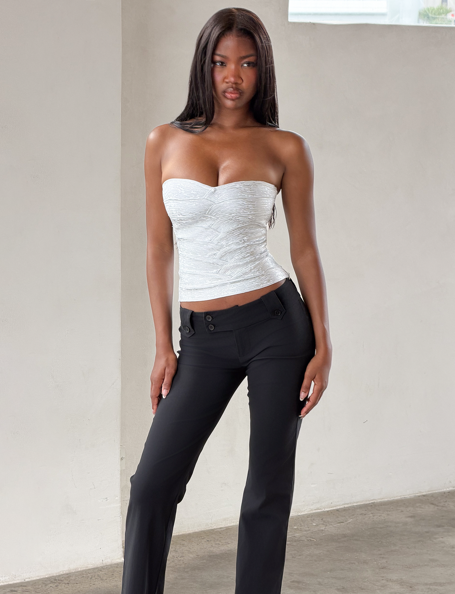 A woman with long straight hair wears the TIGERMIST NIYA BANDAGE TOP in Liquid Silver and black pants, standing indoors against a light-colored wall with one hand relaxed by her side and the other resting on her thigh.