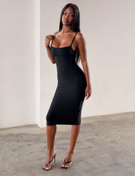 A woman wearing the Tiger Mist USA CL - MIDORI BANDAGE MIDI DRESS and black heeled sandals poses indoors on a concrete floor, confidently touching her shoulder and looking at the camera.