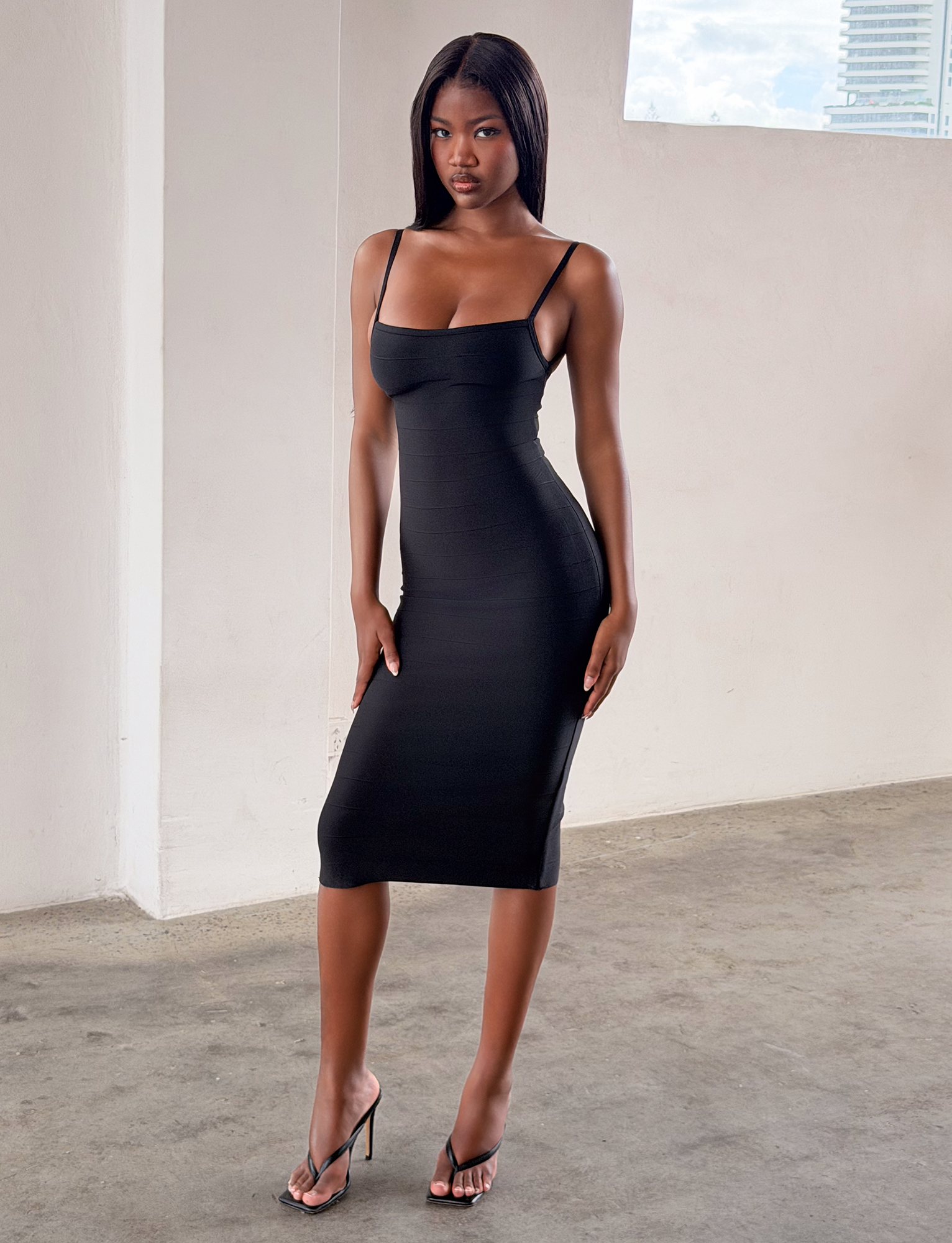 A woman wears the TIGERMIST MIDORI MIDI BANDAGE DRESS in black with black heels, standing indoors on a concrete floor with a neutral wall and large window behind her.