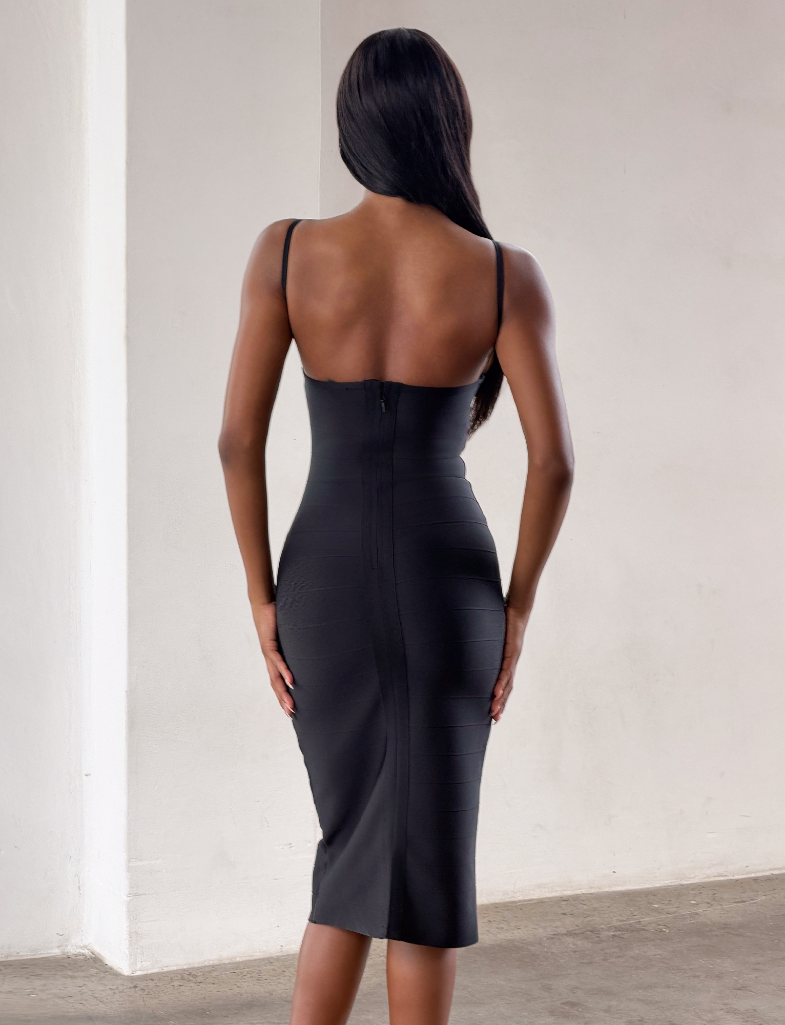 A woman with long dark hair stands facing away in a minimal, light room, wearing the TIGERMIST MIDORI MIDI BANDAGE DRESS in black with thin straps.