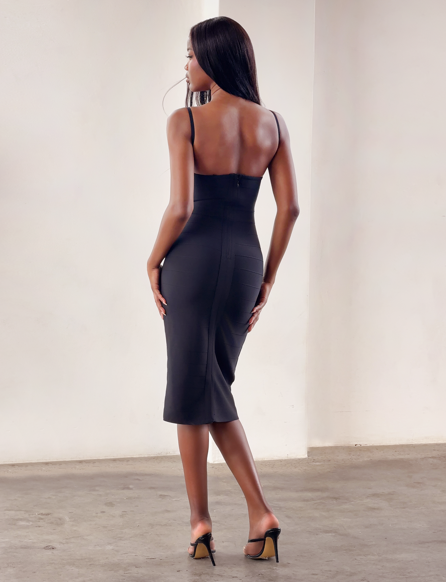 A woman with long hair stands on a concrete floor, facing away from the camera, wearing the TIGERMIST MIDORI MIDI BANDAGE DRESS in black and black high heels against a plain, light-colored background.